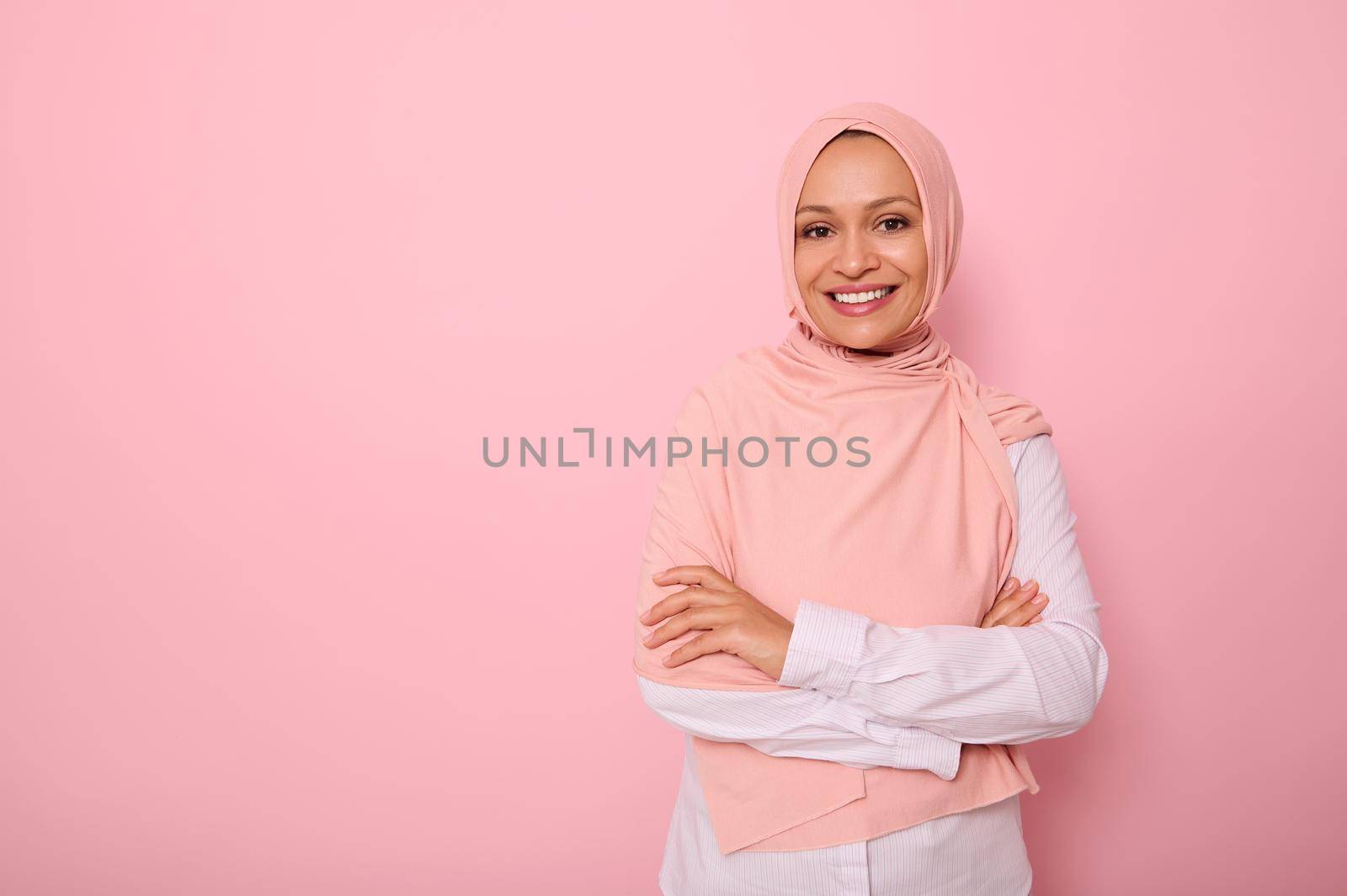 Arab Muslim lady wearing a hijab crossing arms, smiling with beautiful toothy smile, poses on pink background with copy space looking at camera. Concept of successful confident islamic modern woman