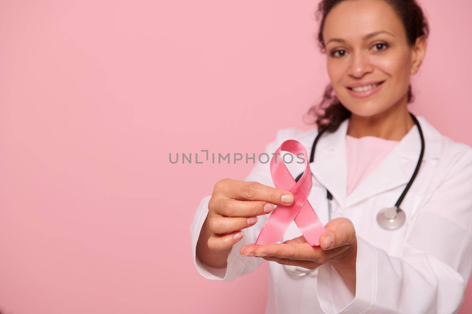 Soft focus on Breast Cancer Awareness Ribbon, in hands of blurred mixed race woman doctor in white medical coat, isolated on pink background, copy space. 1 st October, World Day of fight breast cancer