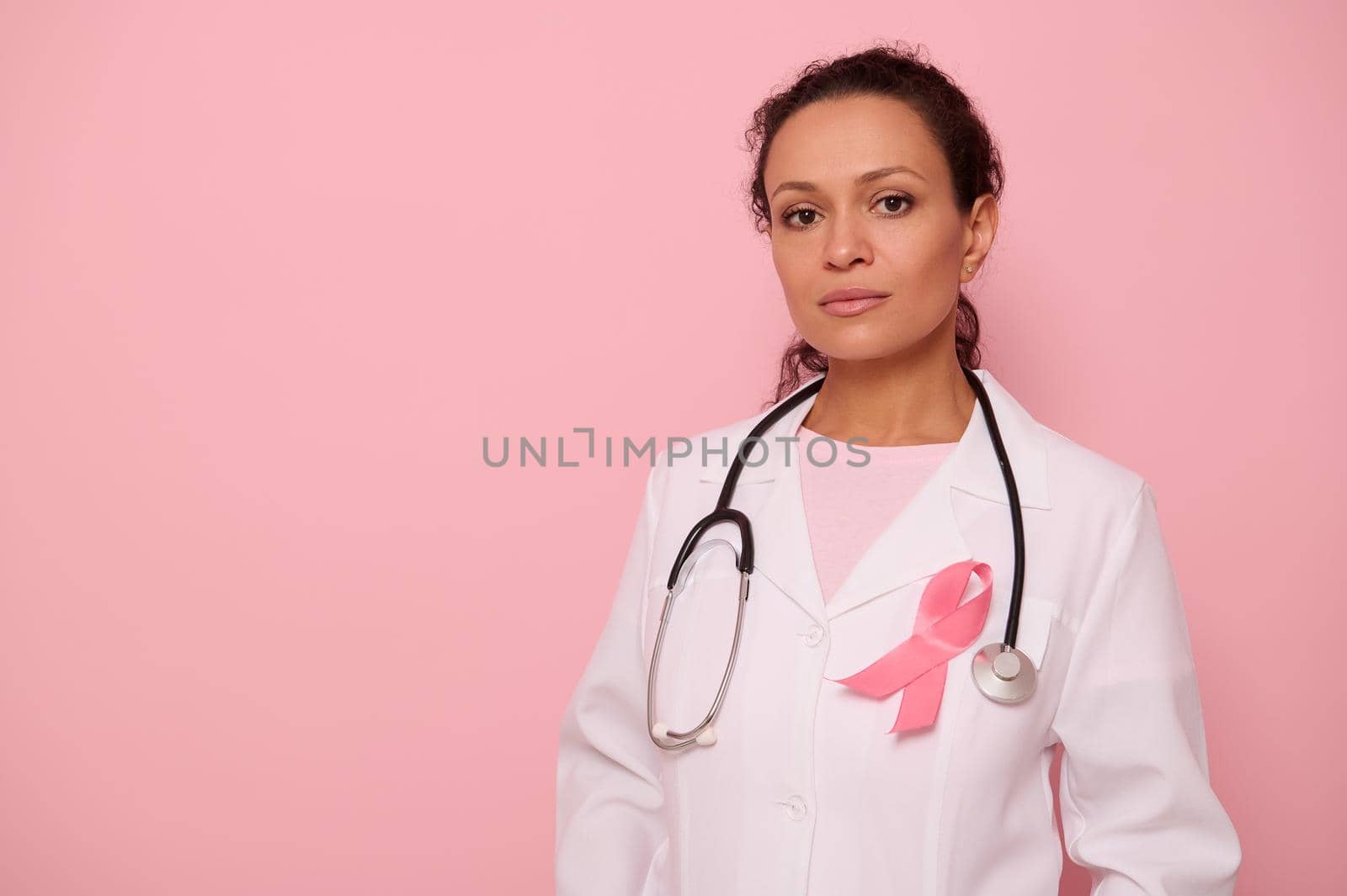 Confident portrait of a mixed race female doctor in medical coat with pink ribbon, and stethoscope around neck, looking at camera, isolated on colored background, copy space. Breast Cancer Day concept