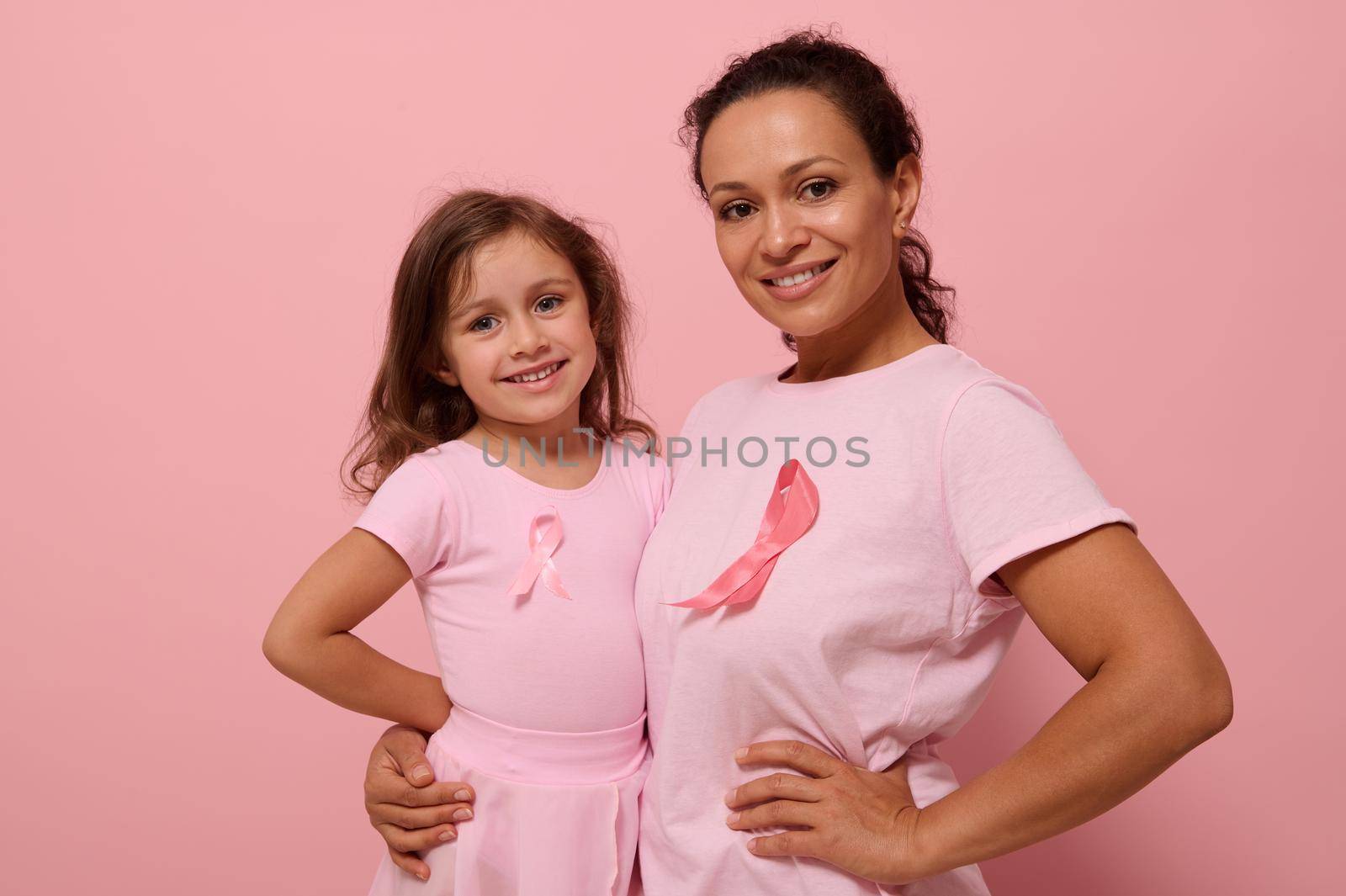 Isolated portrait on colored background with copy space of mixed race woman hugging her daughter, wearing pink clothes and breast cancer awareness ribbon showing solidarity with cancer survivors