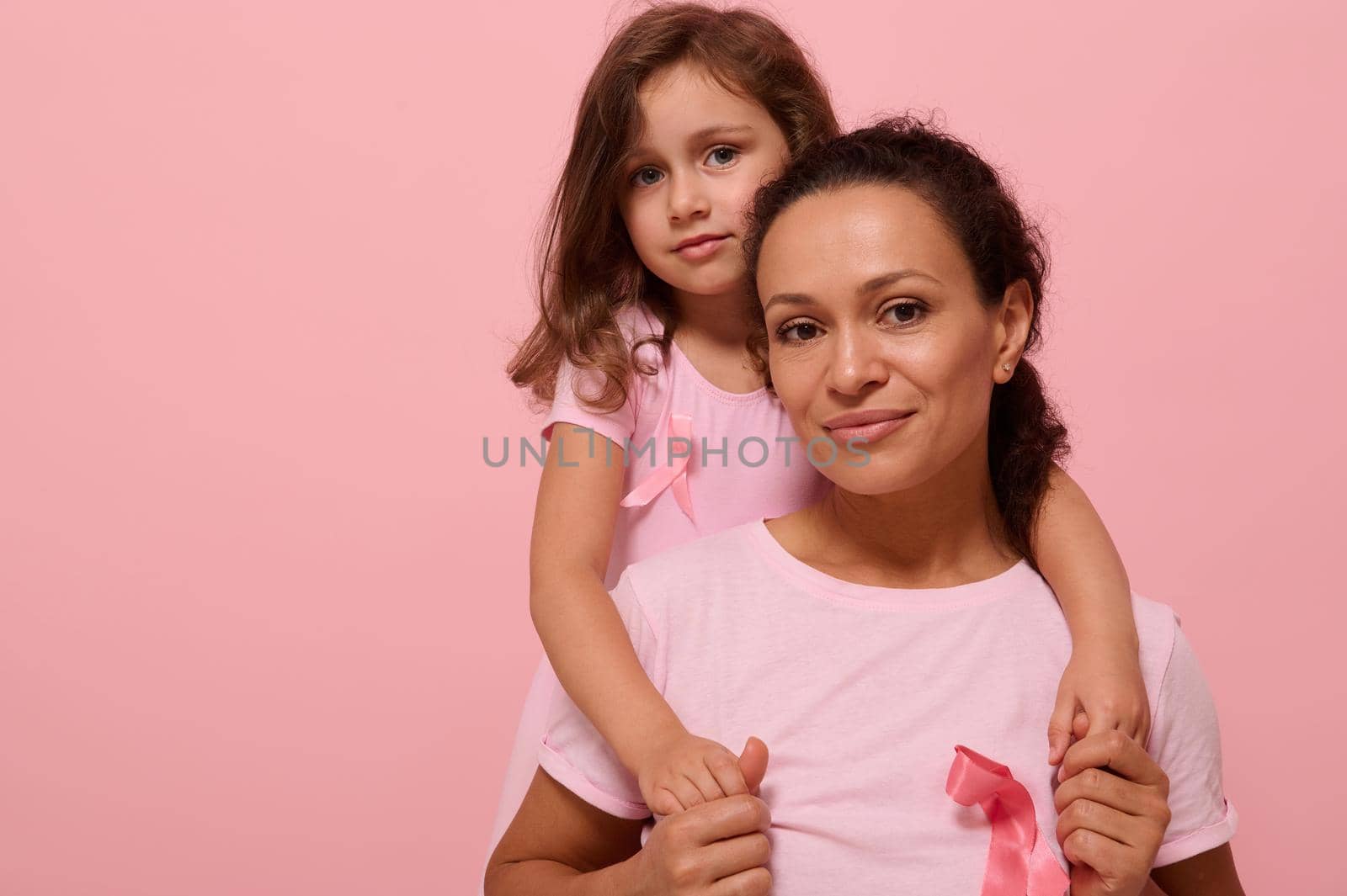 Woman and girl in pink attire with Breast Cancer Awareness ribbon, daughter hugging her mother, holding by hands, looking at camera, supporting cancer patients. Pink background with copy space.
