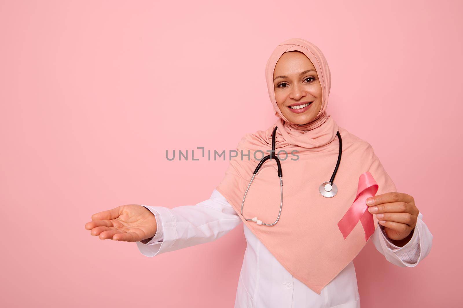 Breast Cancer Awareness campaign, copy space. Friendly Middle Eastern ethnicity Muslim woman in pink hijab and stethoscope around neck, holding pink ribbon and showing with hand on colored background