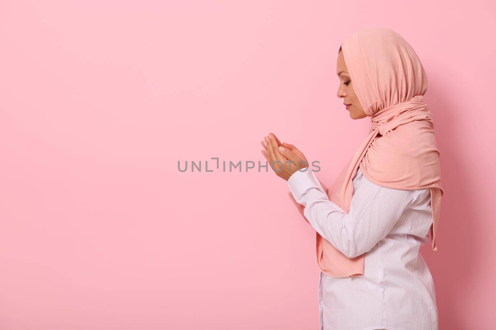 Side portrait of a beautiful and serene Muslim Arab woman in pink hijab and strict outfit with palms folded together at face level performing namaz, isolated on colored background with space for text