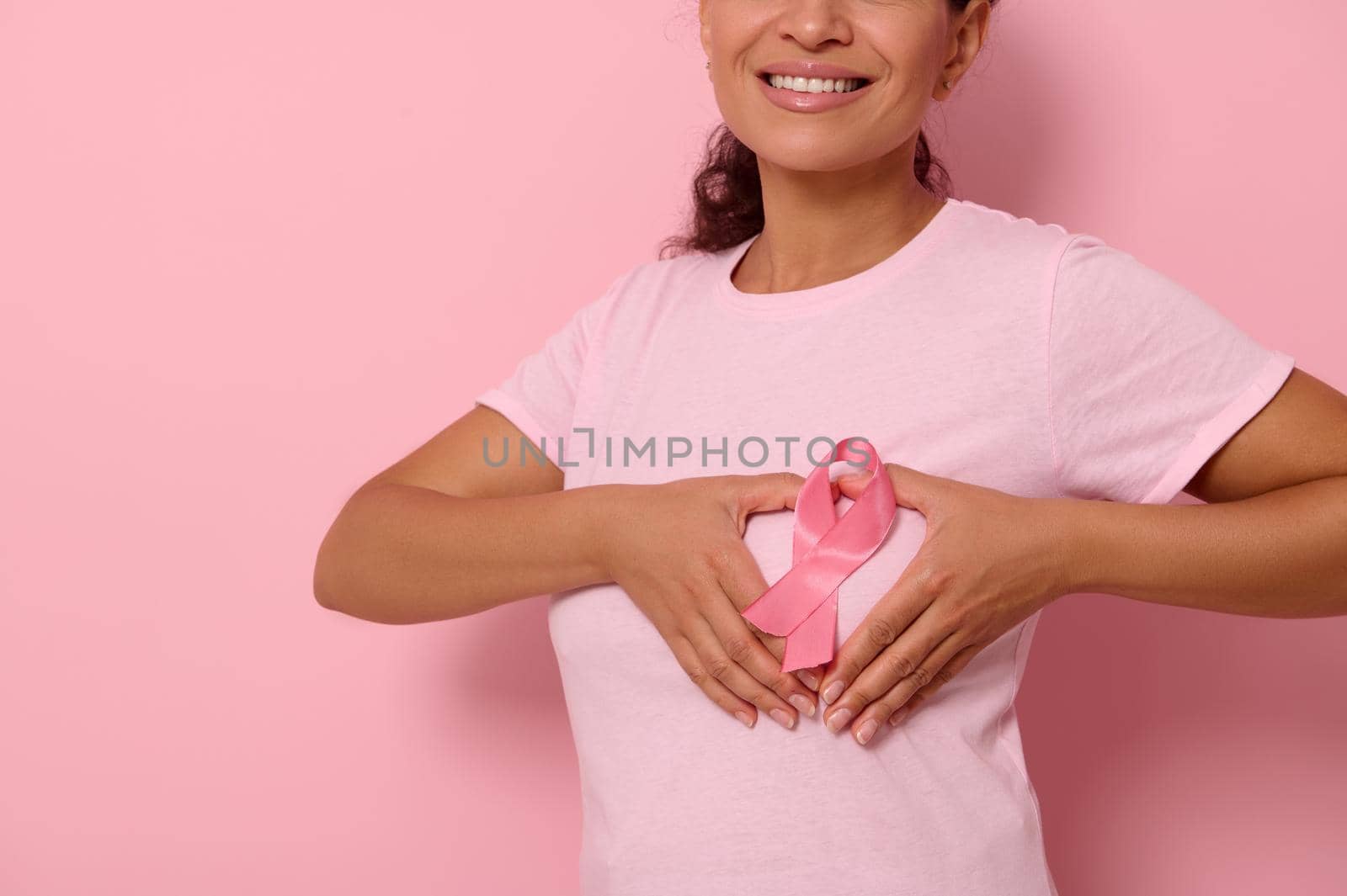 Cropped portrait on colored background of smiling woman in pink t-shirt, putting hands on chest in shape of heart with a pink satin ribbon in the center. World Cancer Awareness Day, fighting cancer
