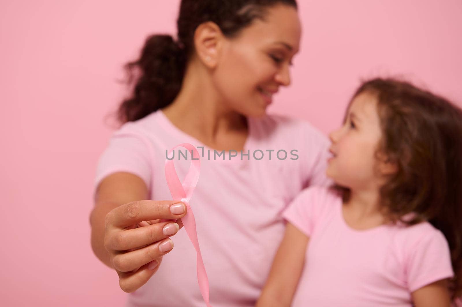 Focus on pink ribbon in the hands of blurred woman and girl wearing pink ribbons and t-shirts - banner for Breast Cancer Awareness Day. Motivational slogan to fight cancer. Copy space