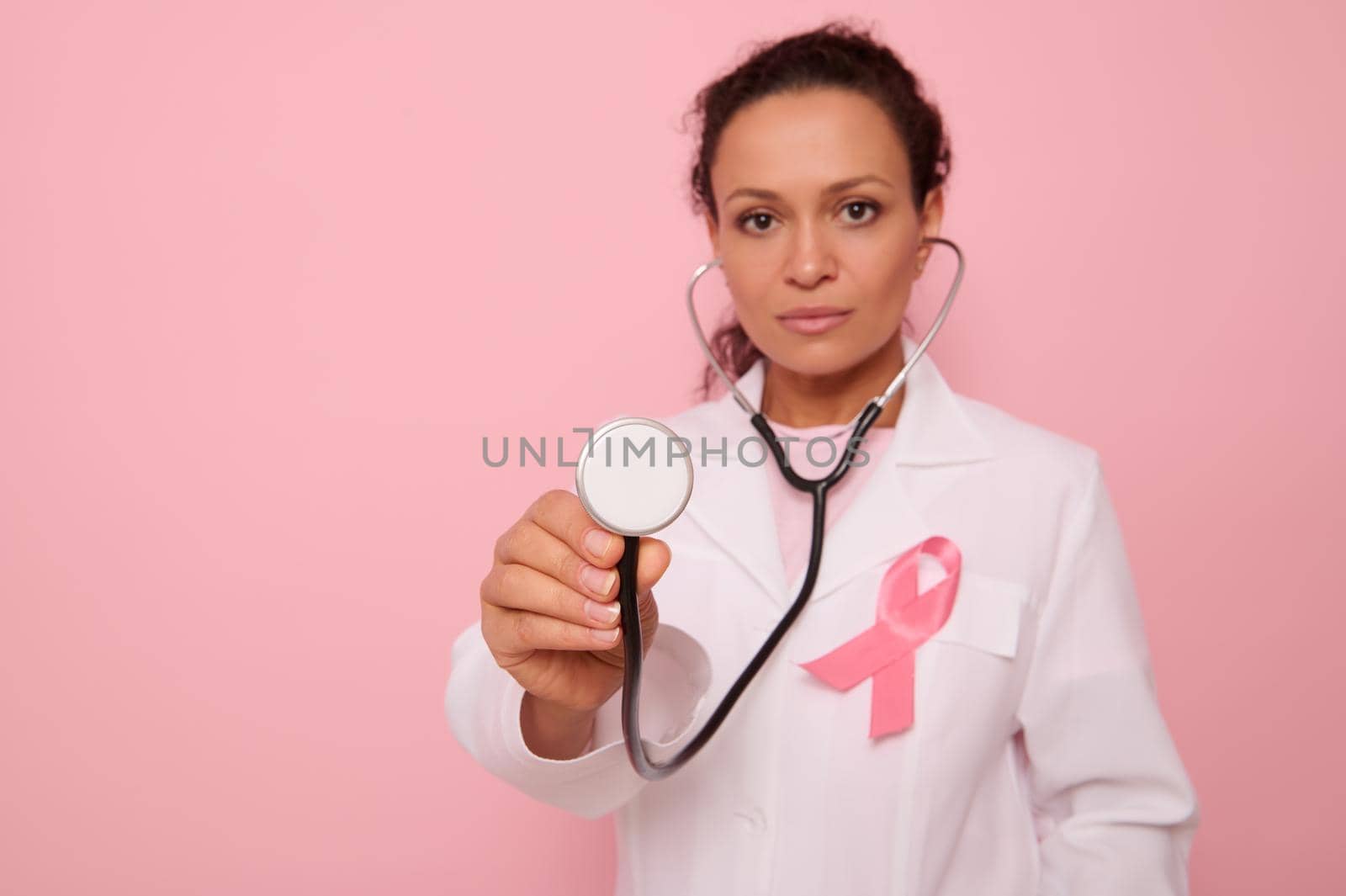 Focus on phonendoscope in the hands of beautiful mixed race Hispanic doctor in medical coat with pink satin ribbon, symbol of Global Breast Cancer awareness Day, Pink October . Woman's health concept