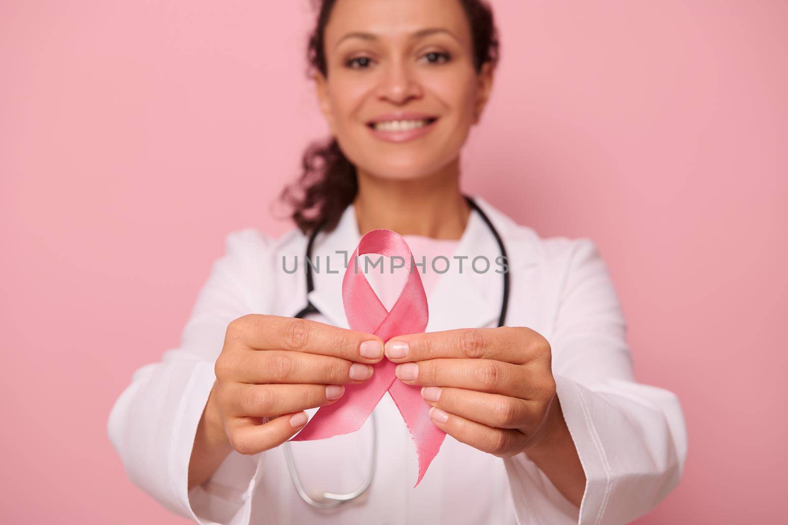Soft focus on Breast Cancer Awareness Pink Ribbon, in hands of blurred mixed race female doctor in white medical coat, isolated on colored background. 1 st October, World Day of fight breast cancer by artgf