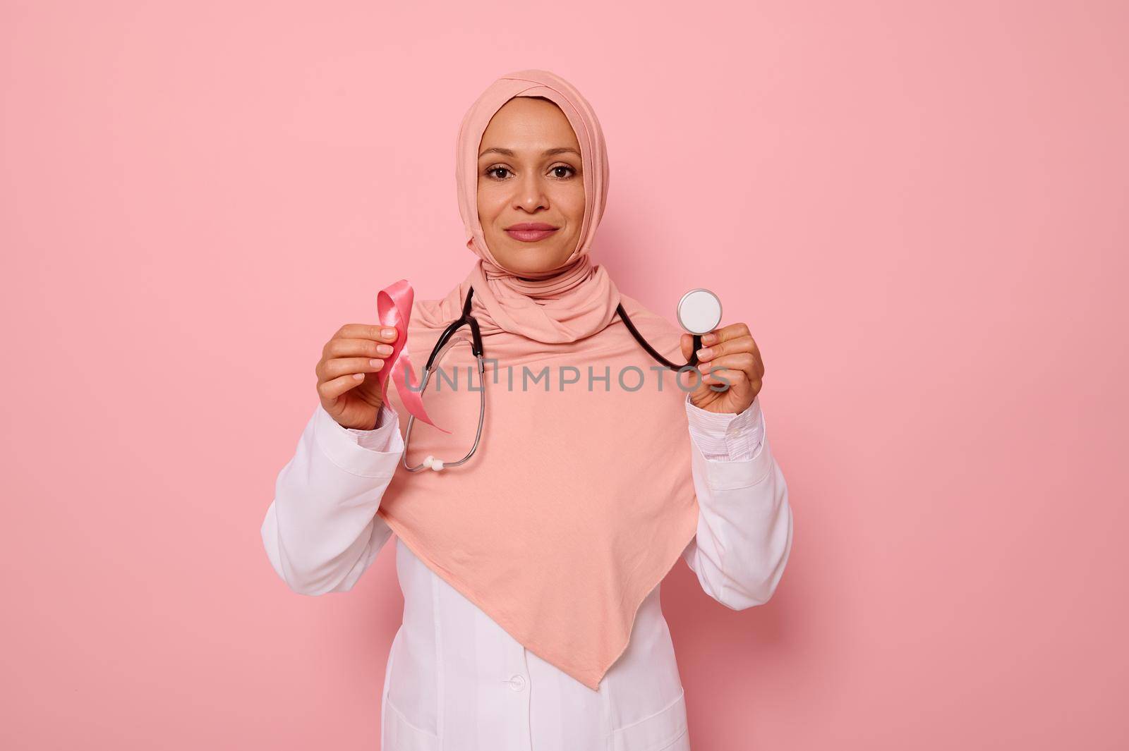 Portrait of Muslim Arab woman doctor with covered head in pink hijab, posing on colored background with a pink ribbon and stethoscope in hands, supporting cancer patients and survivors, 1 st October