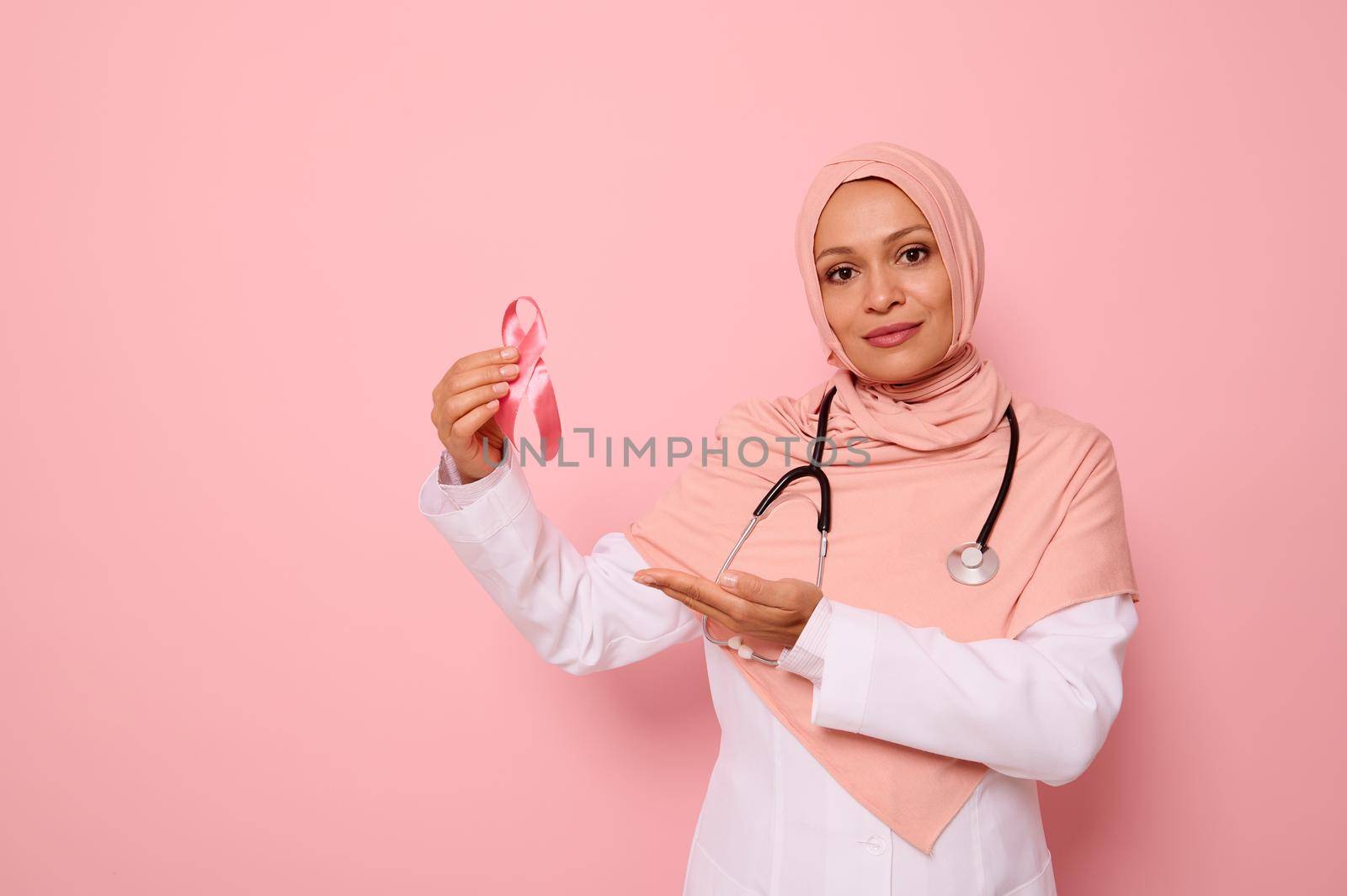 Confident Arab Muslim woman doctor in pink hijab and medical coat, showing pink Cancer Awareness ribbon, looking at camera. Pink background, copy space. International Day of fight oncological illness