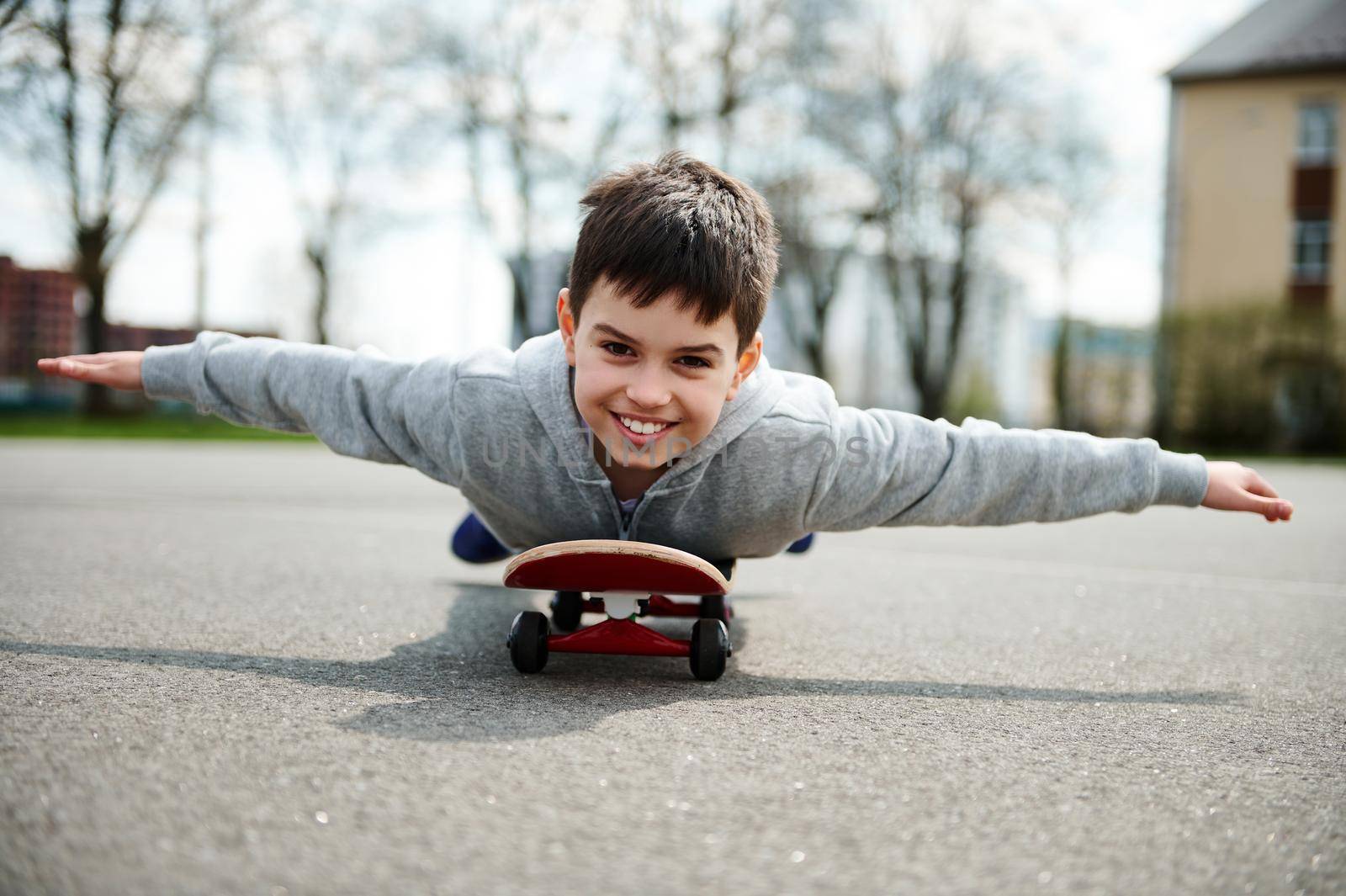 A cute boy lying with his belly on a skateboard with his arms outstretched like wings and imagining a flight by artgf