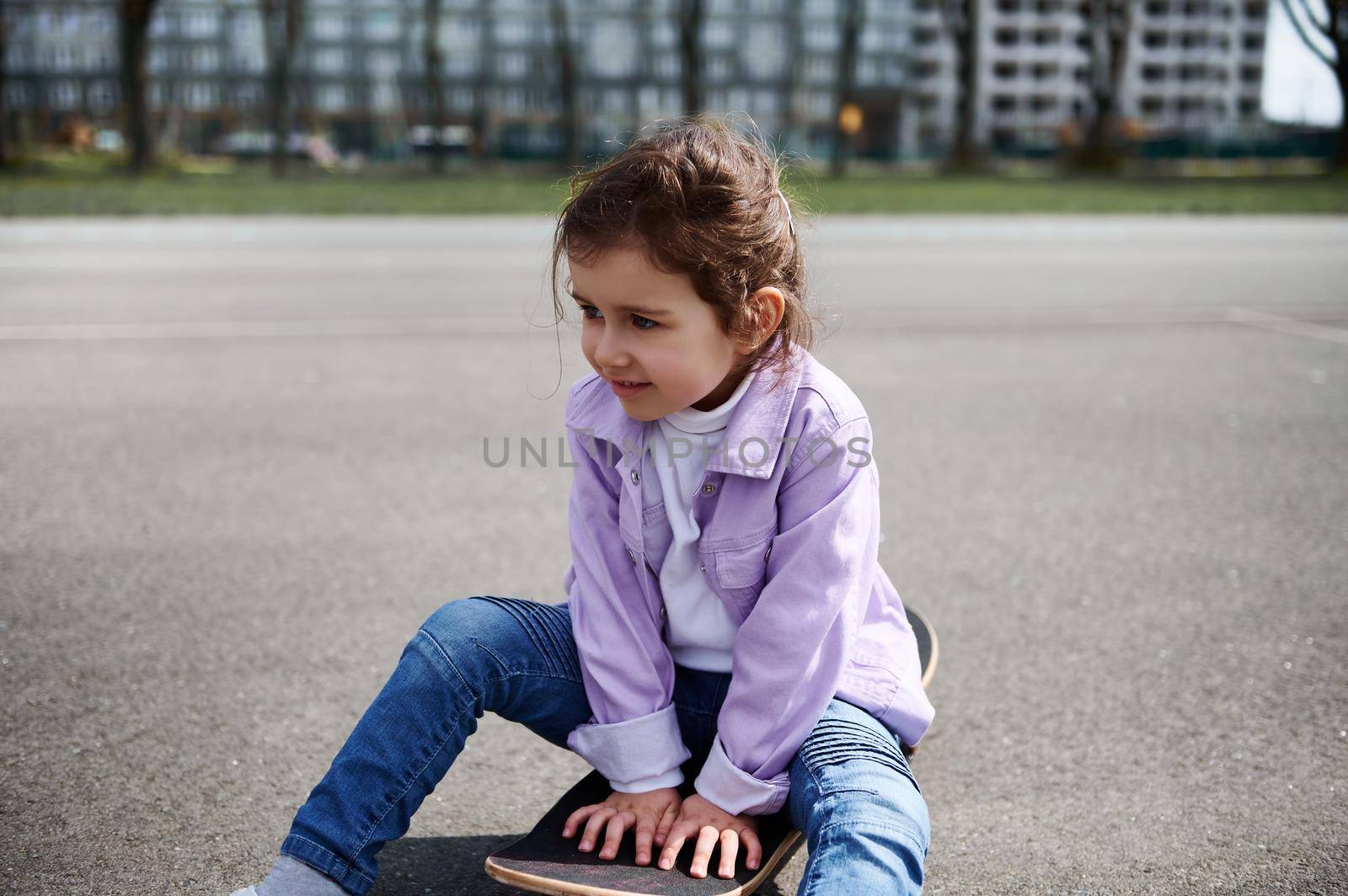 Adorable female child sitting on wooden skateboard against blurry buildings background by artgf