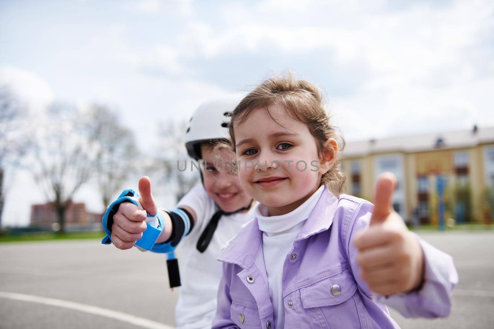 Face portrait of adorable children, boy in skateboard helmet and girl sitting next to each other and showing thumb up, on the street background