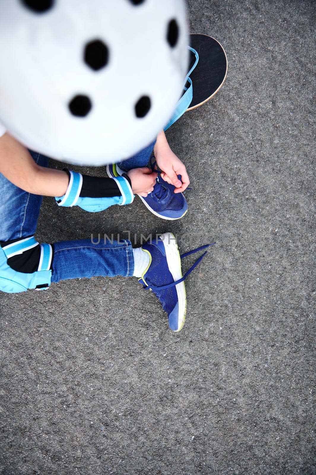 High angle view of unrecognizable boy in safety helmet for skateboard tying shoelaces sitting on asphalt of playground