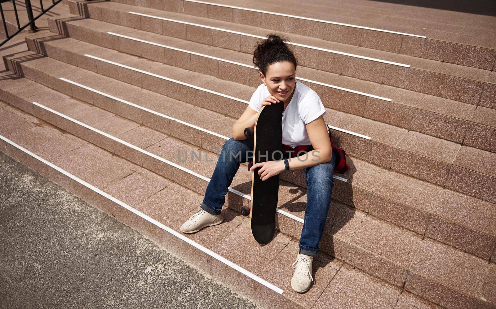 High angle view of a happy smiling mixed race woman with skateboard sitting on steps outdoors