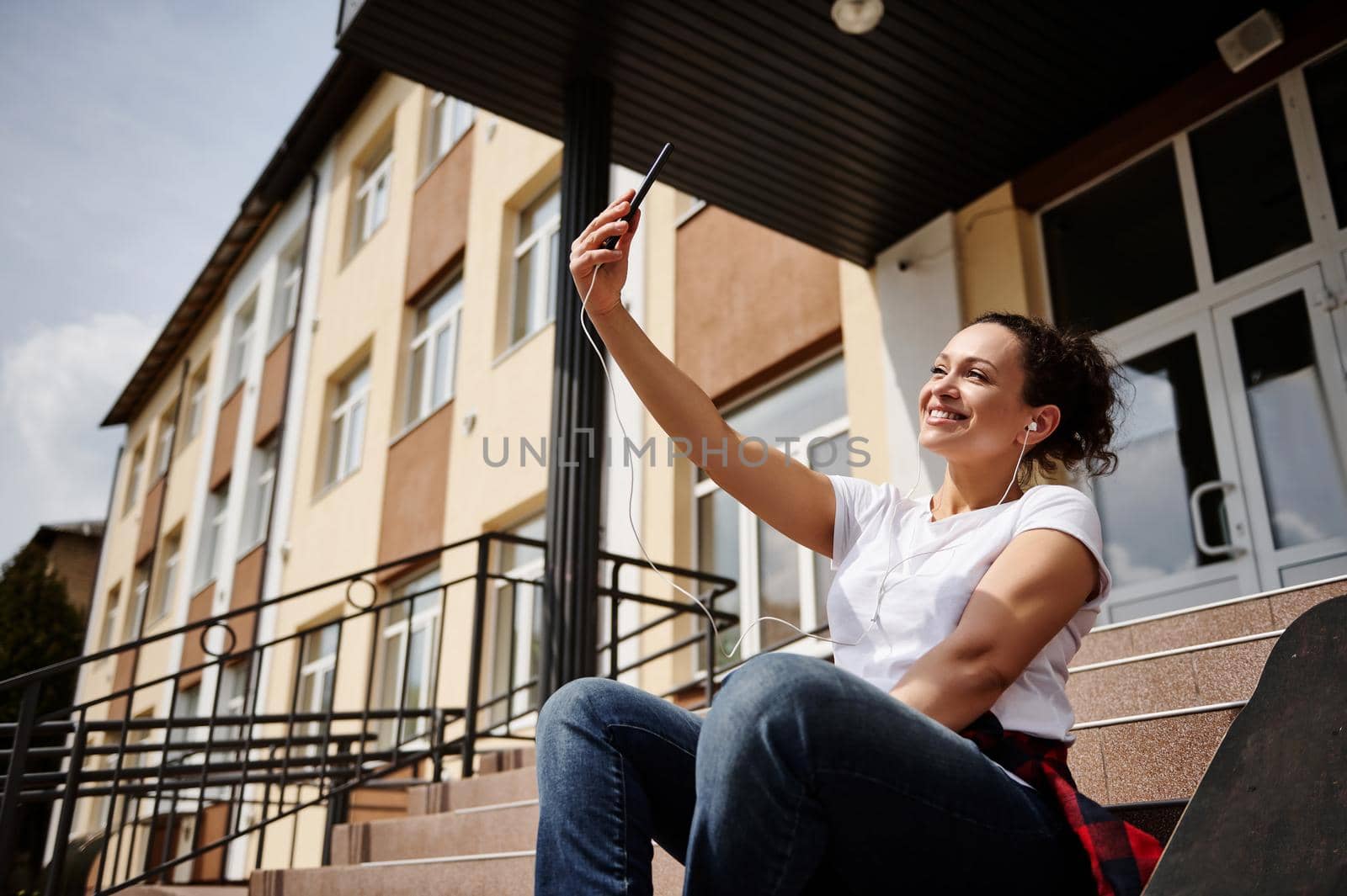 Smiling happy African American woman with headphones in casual clothing making selfie while sitting on steps on the background of yellow building