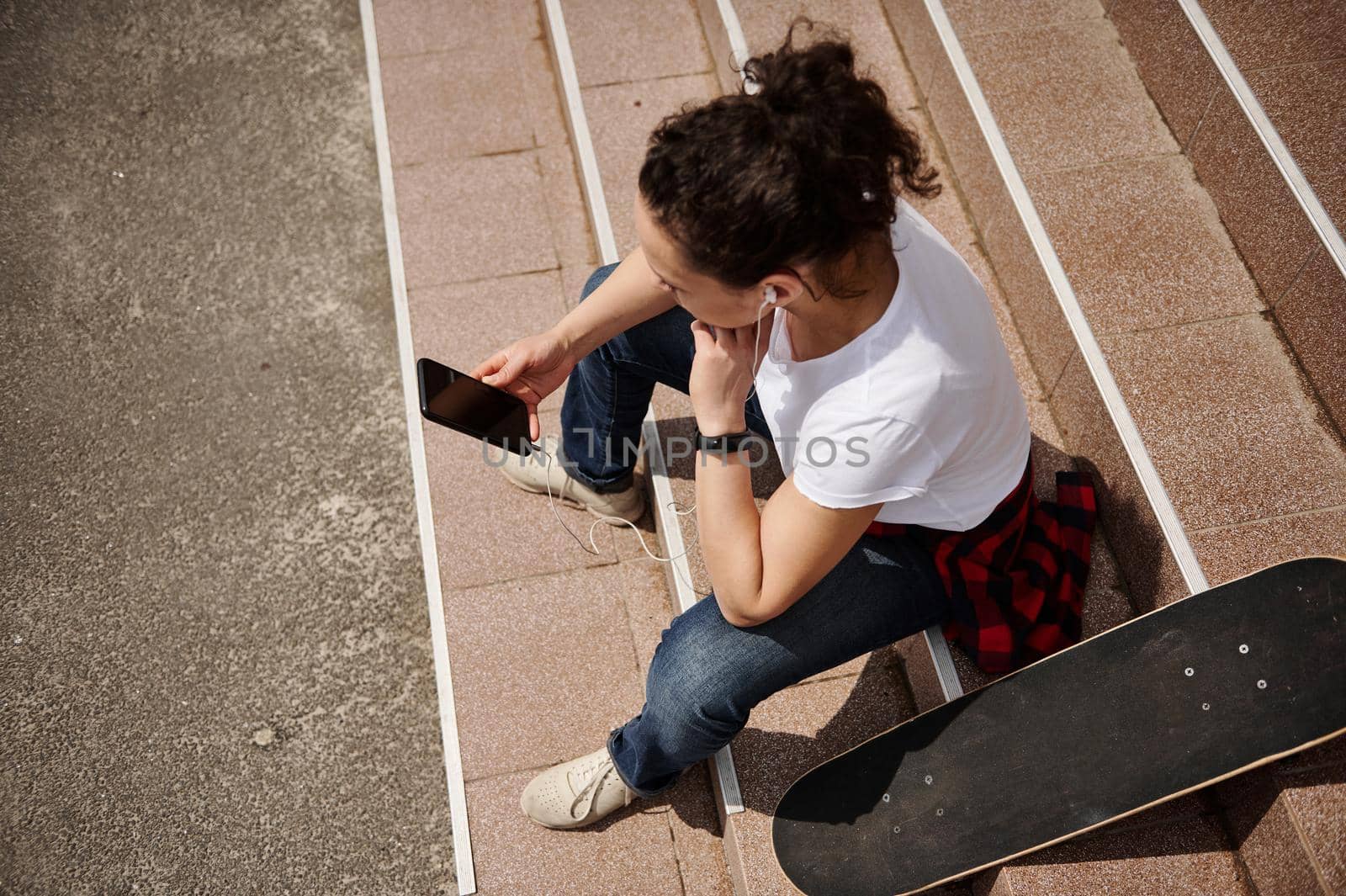 High angle view of a young woman with skateboard sitting on steps and holding a mobile phone by artgf