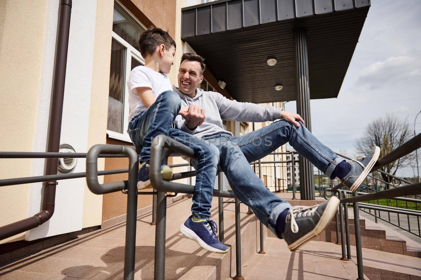 Handsome caucasian man laughing with his adorable son while sitting on a handrail outdoors