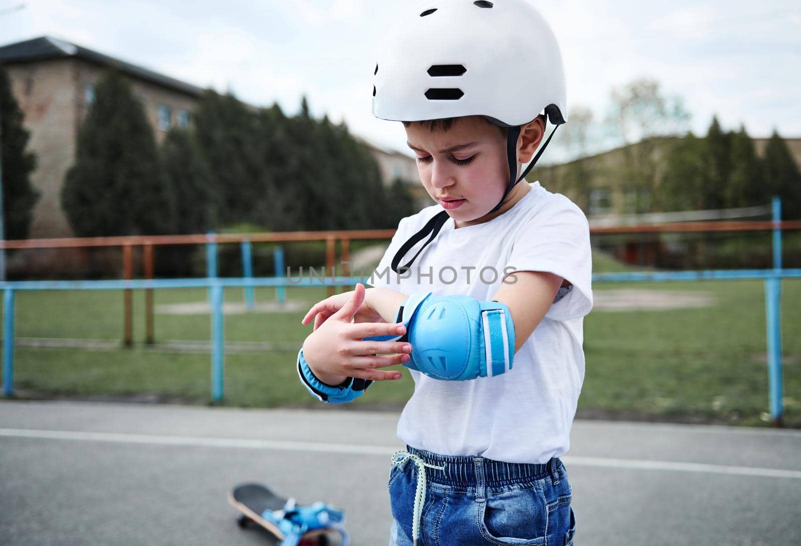 Portraot of a handsome boy skateboarder wearing safety helmet putting on protective armrests before skating by artgf
