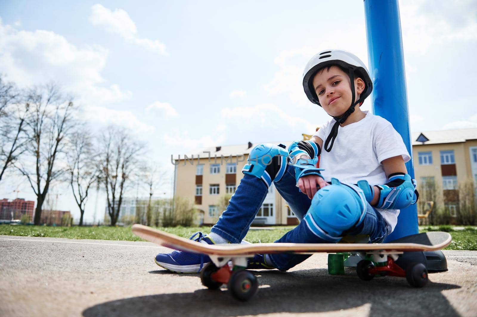 Bottom view of smiling boy in protective gear of skateboarder sitting on skateboard and looking at camera against the background of a yellow building