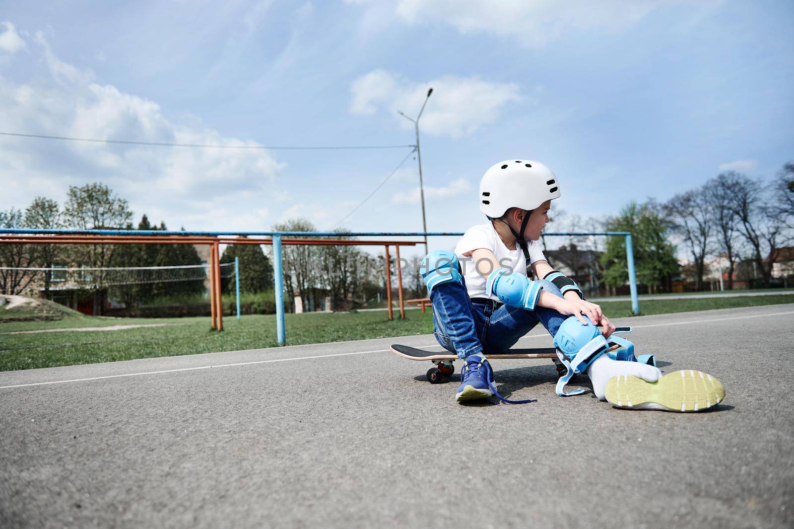 Child boy skateboarder looking away while sitting on a skateboard and putting on protective equipment. Concept of leisure activity, sport, extreme, hobby.