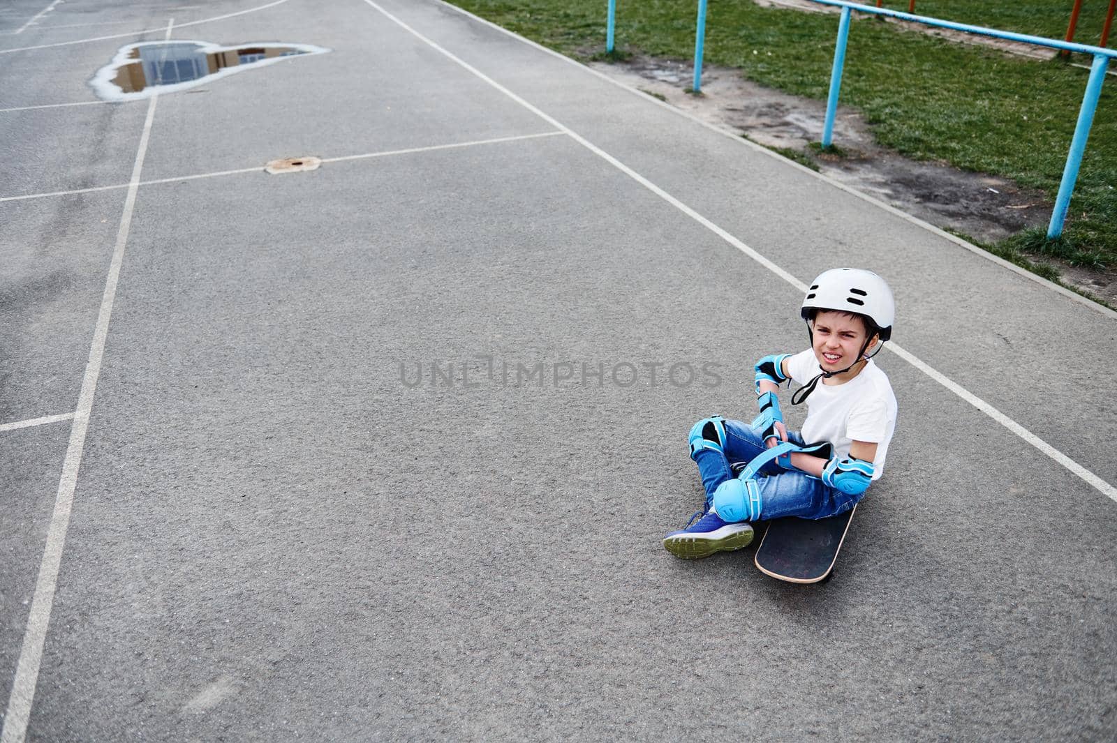 Boy in safety helmet sits on skateboard and focuses on putting on protective gear for skateboarder