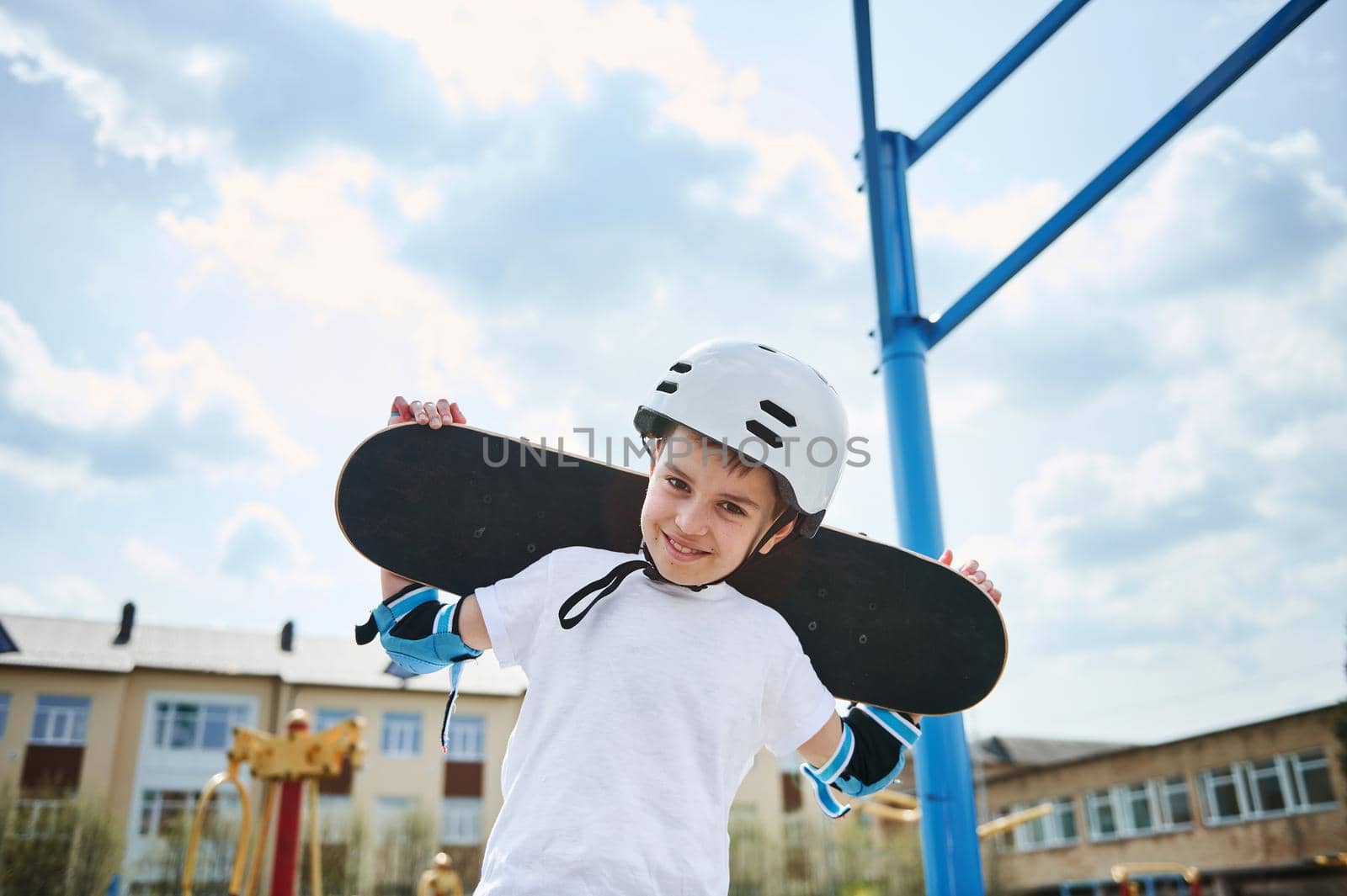Adorable boy in a protective helmet and equipment posing with a skateboard on his shoulders behind his head by artgf