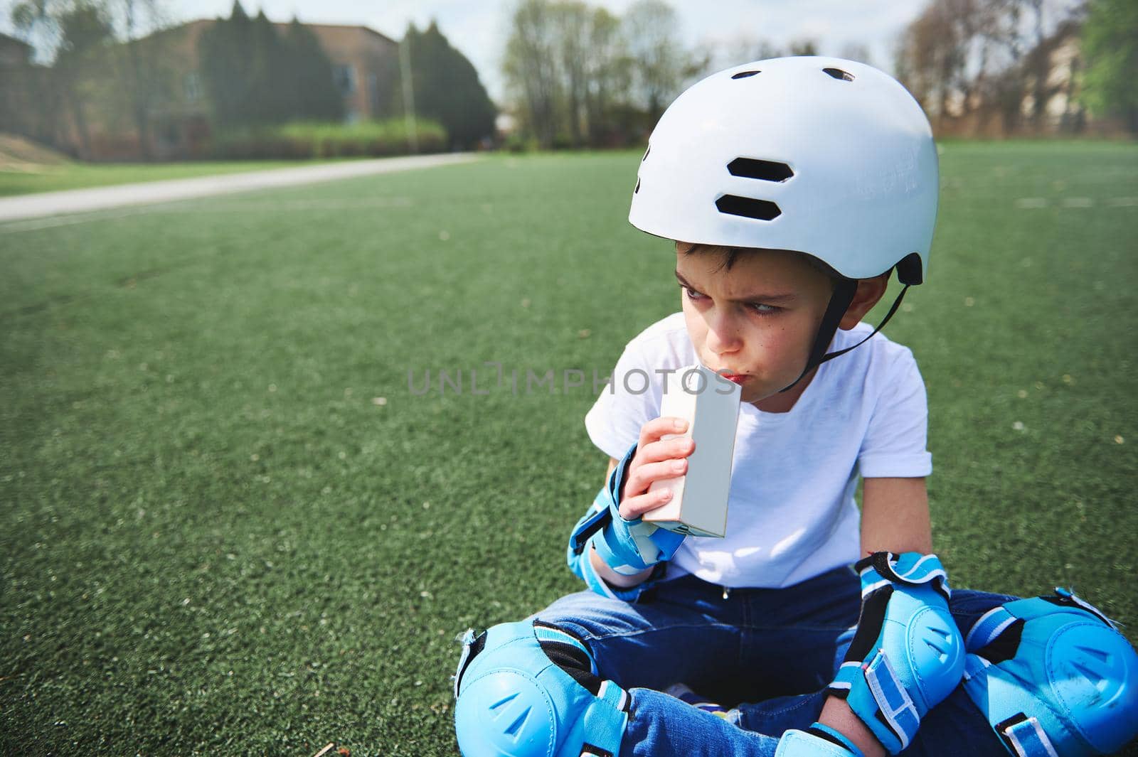 Cute boy in a skateboard helmet and protective equipment resting sitting on a green grass and drinking juice by artgf