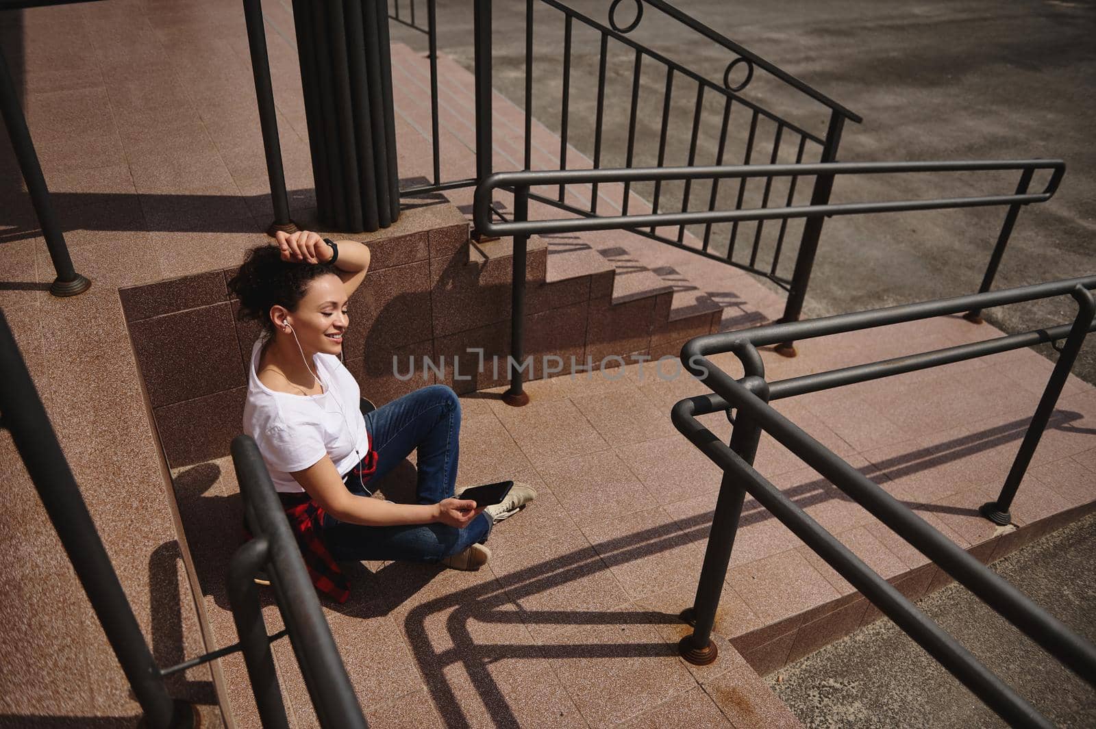 Smiling African American woman sitting on skateboard with headphones and smartphone in her hand and enjoying beautiful warm sunny day