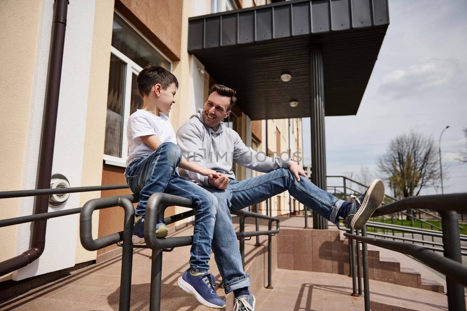 Happy young father sitting on the handrail and happily communicating with his son on a warm spring day. Ideal family relationships