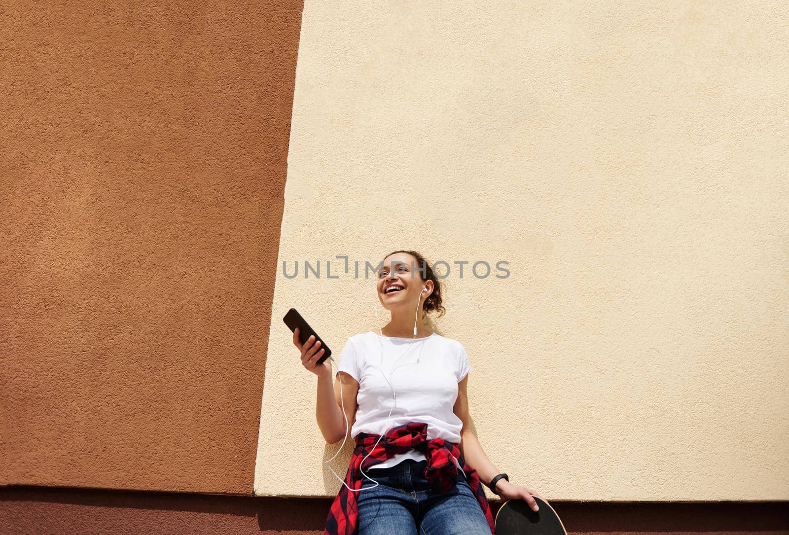 Young smiling brunette woman leaning against colored wall posing with skateboard and mobile phone in hands