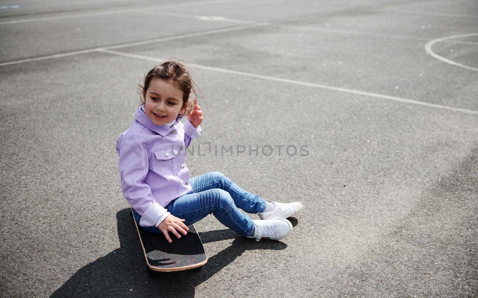 Cute little girl sitting on a skateboard on a sports playground on a beautiful sunny day