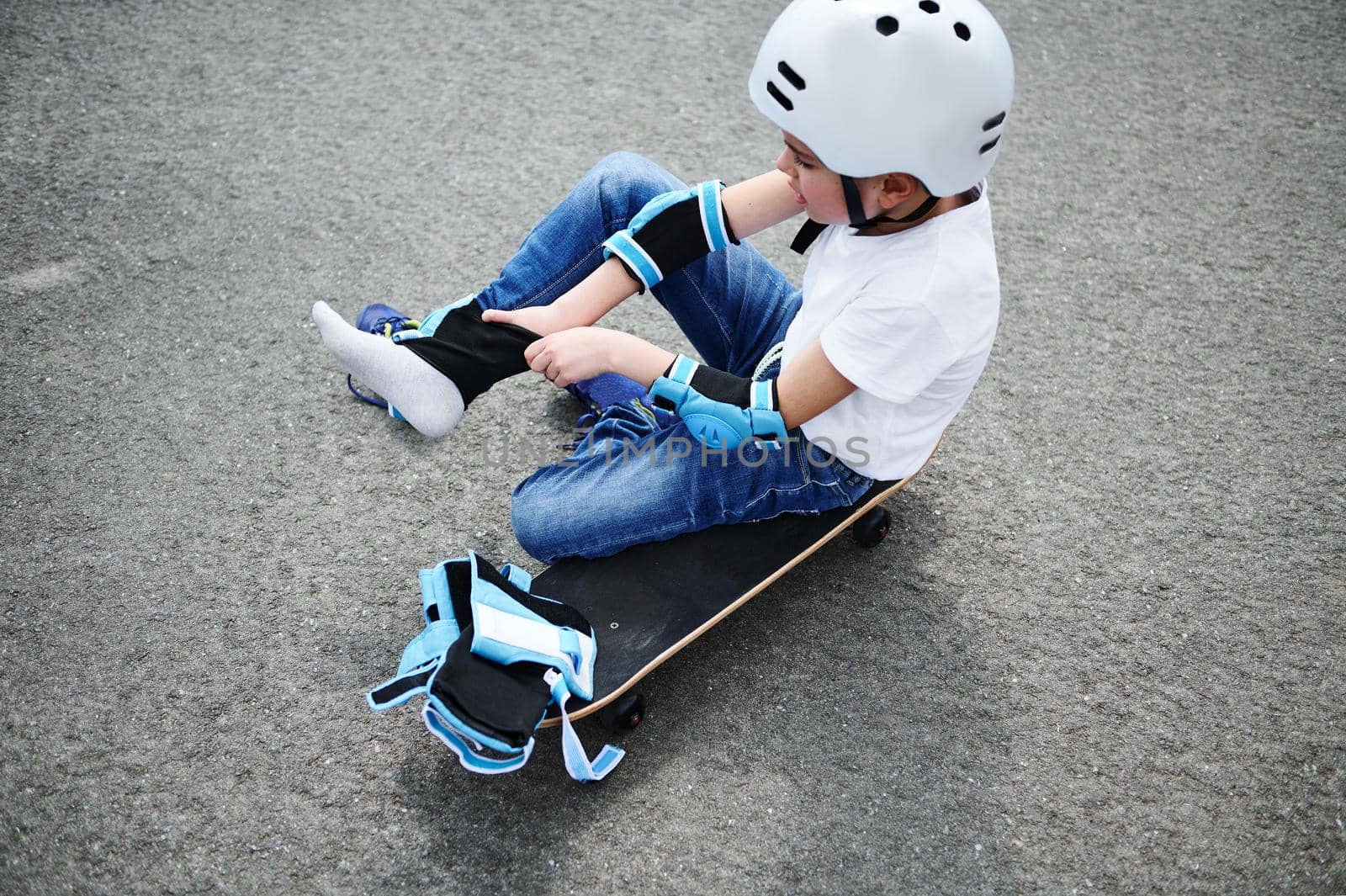 High angle view of a sporty boy in safety helmet sitting on skateboard on playground asphalts and putting on protective knee pads