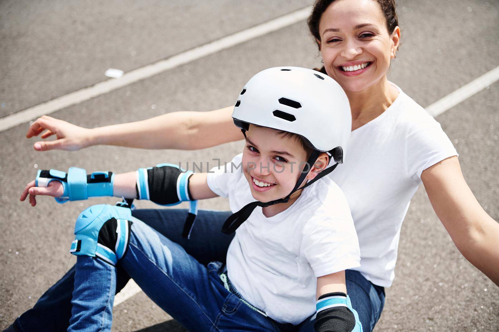 Happy mom and adorable son in protective helmet and equipment enjoying time together sitting on a skateboard by artgf
