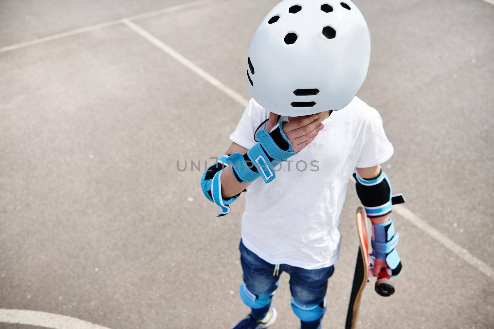 Teenage boy in protective helmet and gears holding a skateboard, looking down and wiping tears from his eyes by artgf