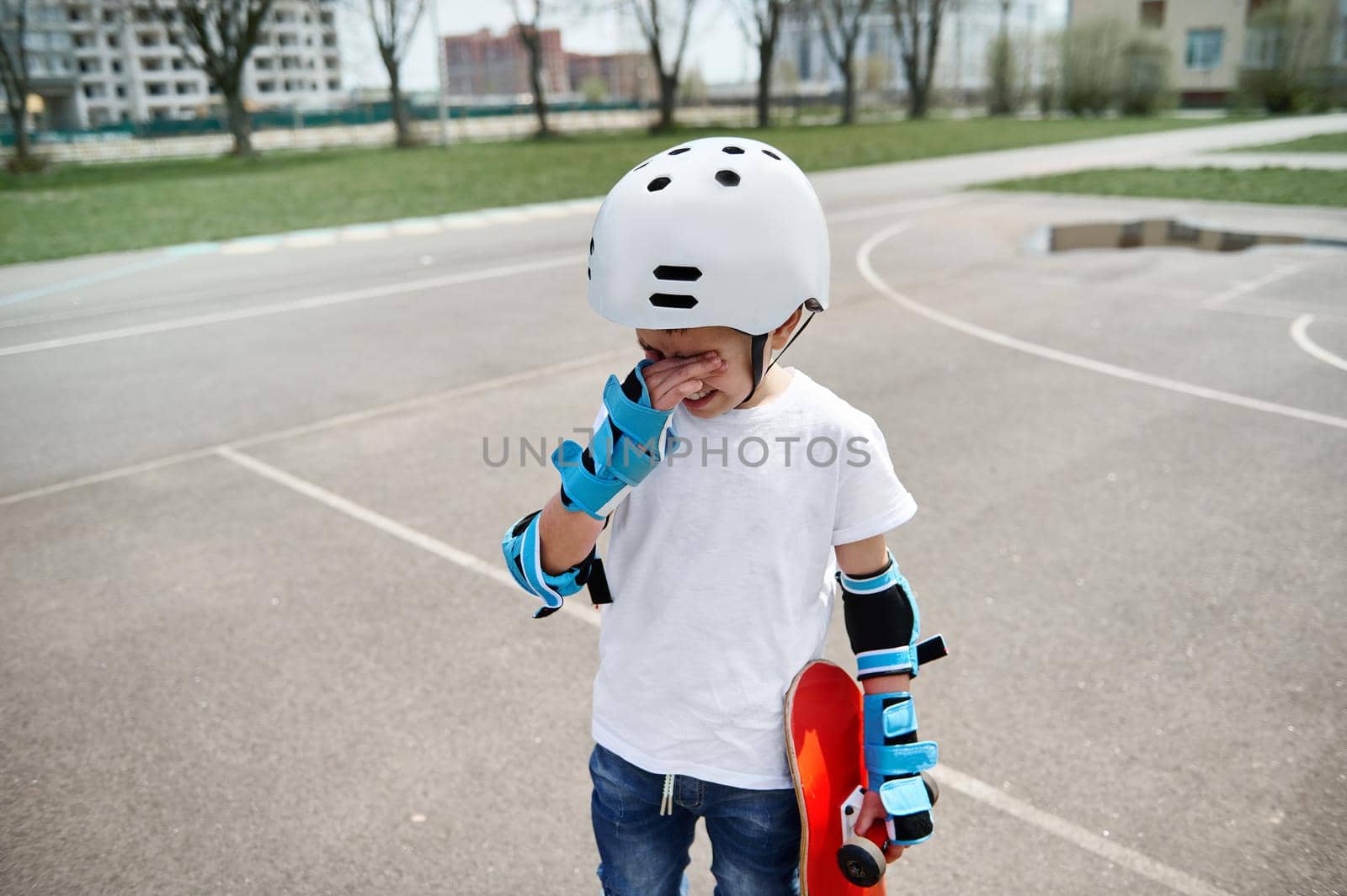 Upset boy skateboarder in protective gear frustratedly wipes tears from his eyes and holds a skateboard in one hand while standing on a sports playground against the background of buildings