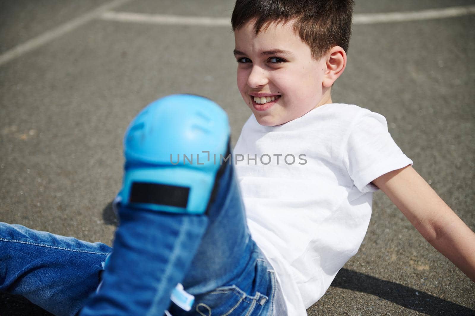 Close-up portrait of a beautiful child in a white t-shirt and protective knee pads sitting on the asphalt outdoors