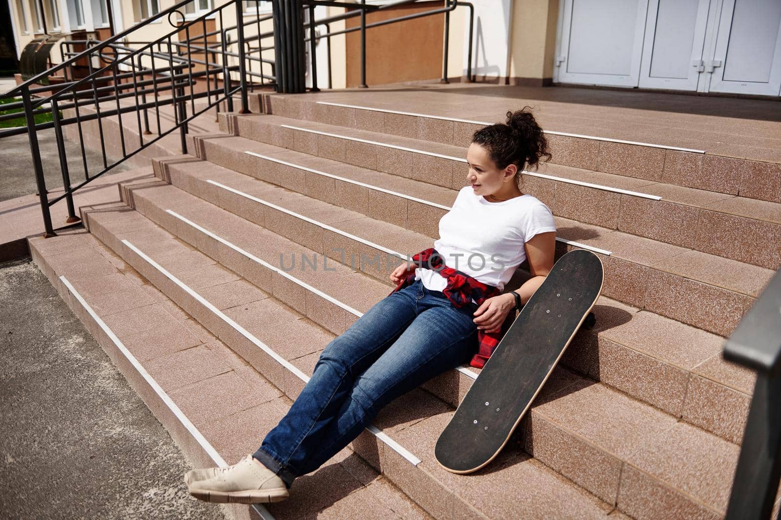 Attractive African American woman in casual clothing posing on stairs with skateboard and enjoying sunny day by artgf