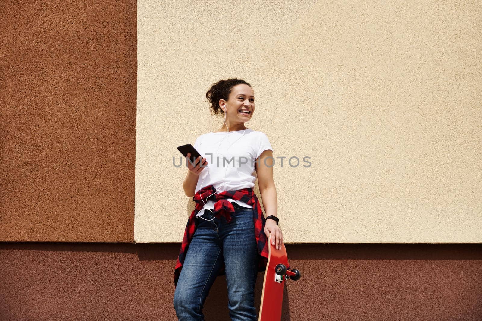 Female skateboarder with wooden red skateboard holding smartphone, enjoying listening to music posing outdoors near wall by artgf