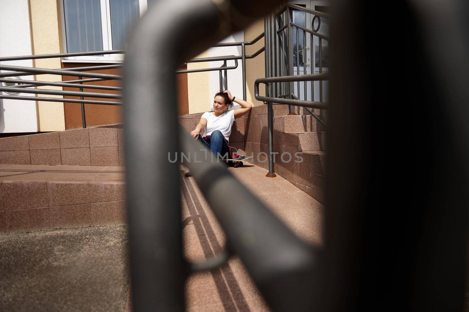 View through the railing of a young woman sitting on skateboard and having rest on a beautiful warm and sunny day by artgf