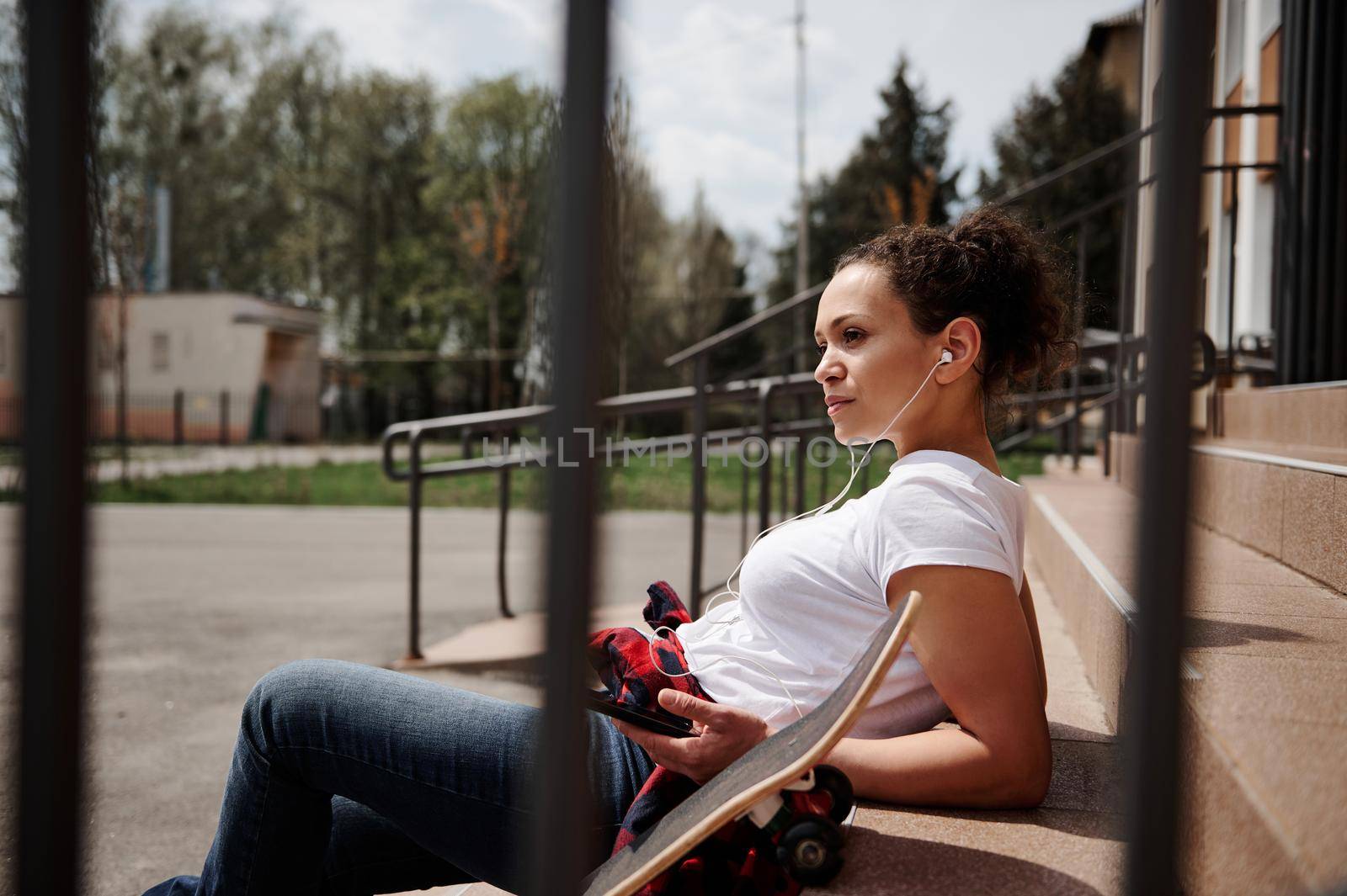 Charming Latin American woman with headphones posing leaning on steps outdoors by artgf