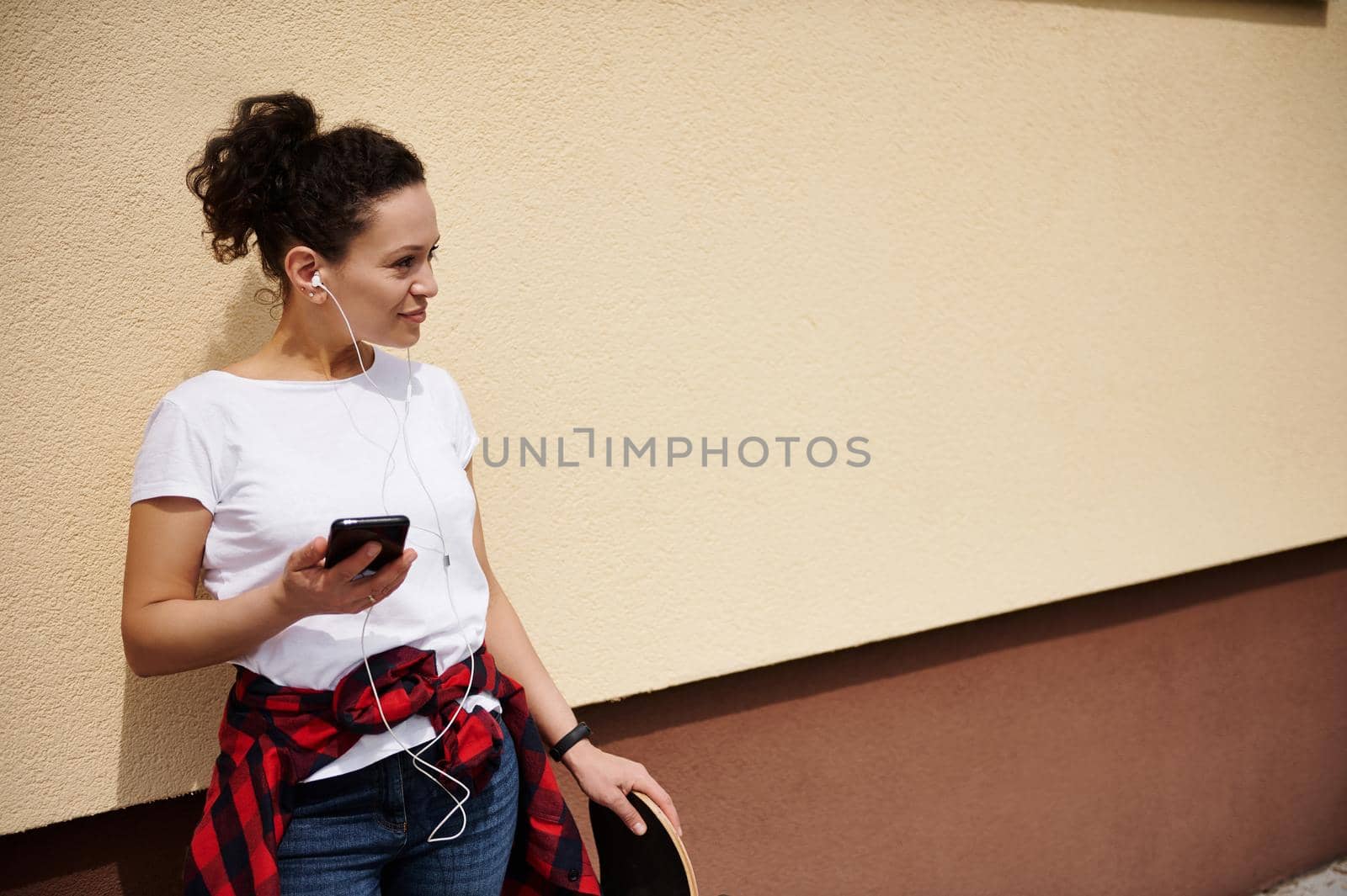 Attractive mixed race young woman with curly hair holding a mobile phone in one hand and a skateboard in the other, looking to the side with her back leaning against a colored wall outdoors