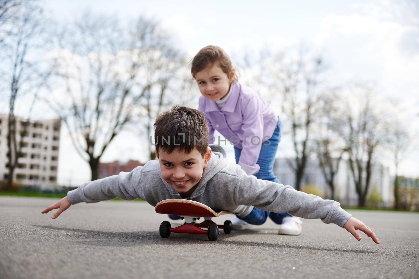 Smiling girl riding her brother on a wooden skateboard enjoying a game together on a sports ground by artgf