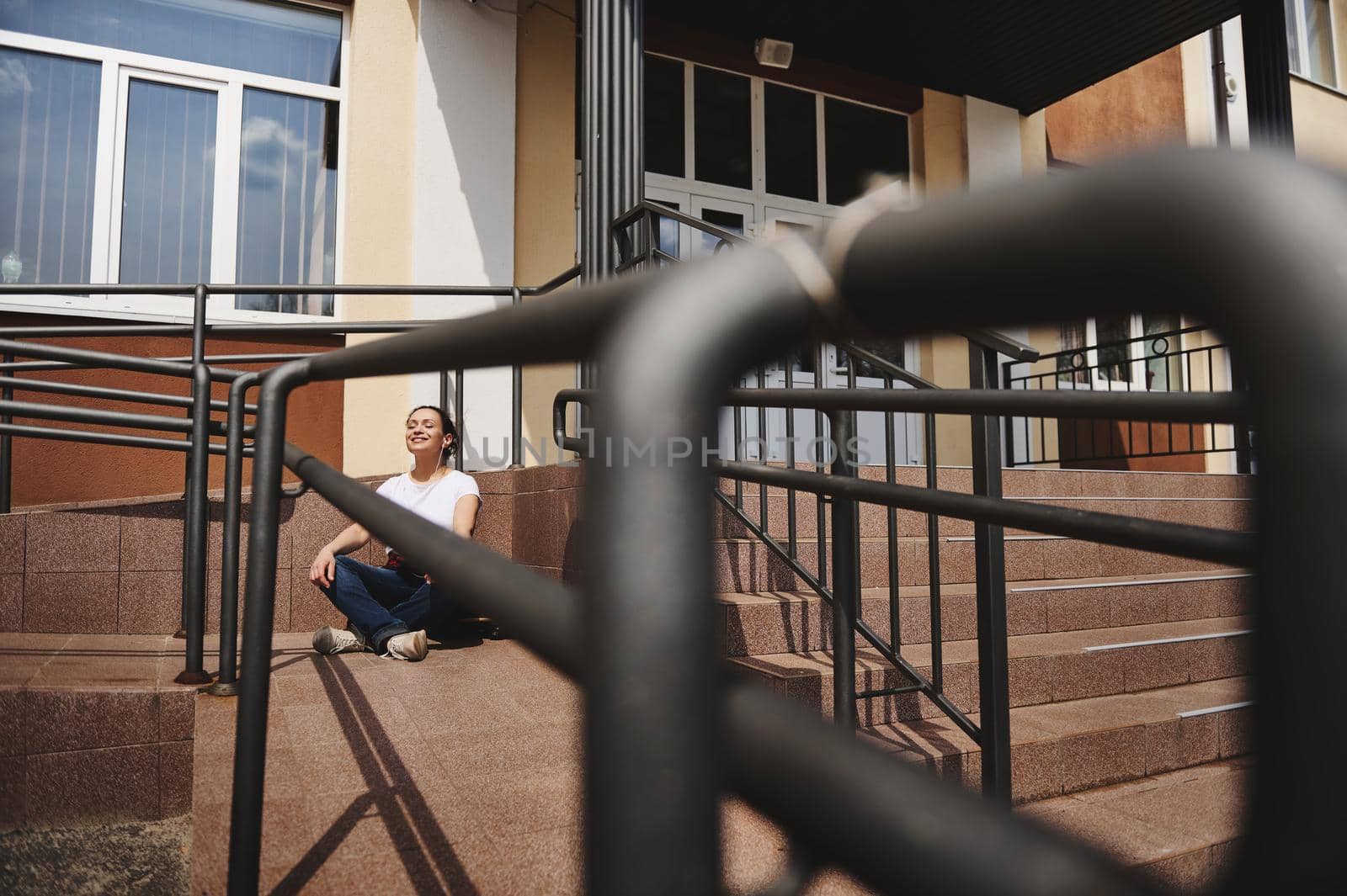 View through the railing of a young Latin American woman sitting on skateboard in lotus pose and having rest on a beautiful warm sunny day