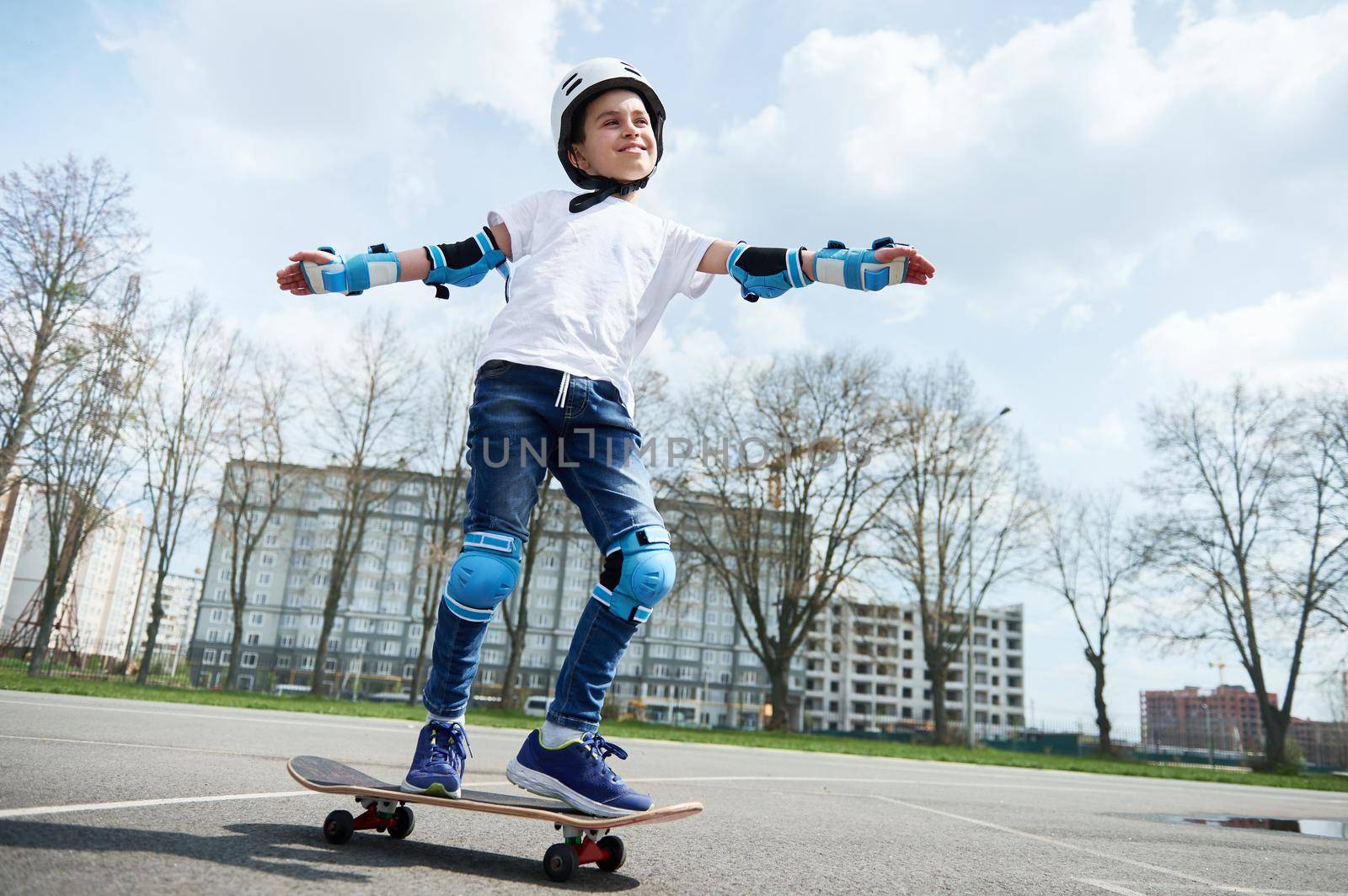 Happy and smiling boy in protective gear and helmet keeps balance while riding a skateboard by artgf