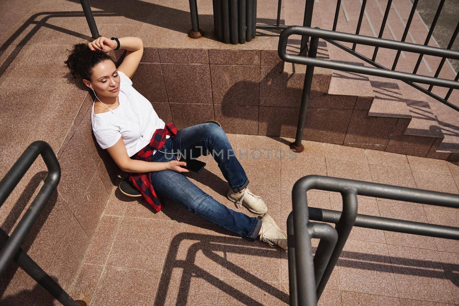 Attractive young woman listening to music on headphones and having rest leaning on steps of unrecognizable building, enjoying warm sunny day