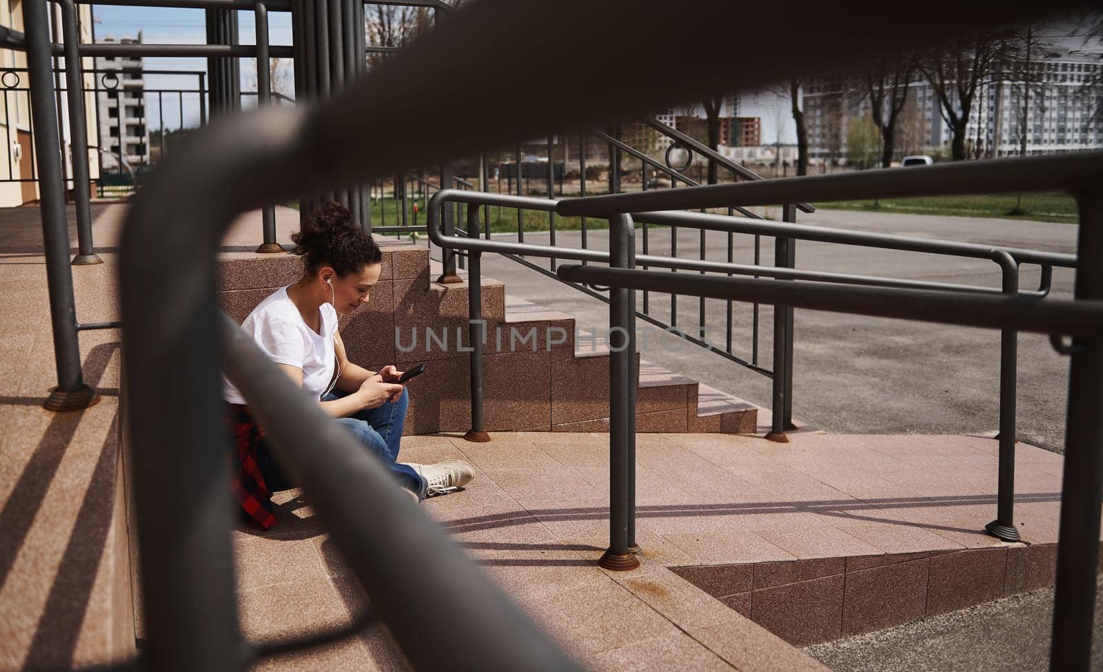 Young woman in headphones using mobile phone while sitting on skateboard on background of steps by artgf