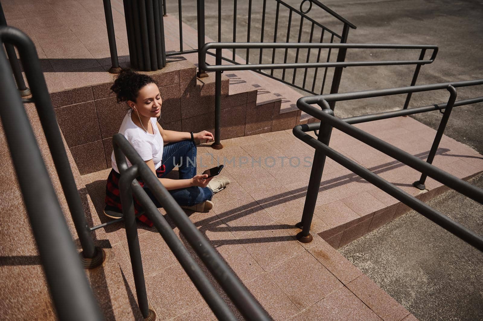 Young Latin American woman in headphones using mobile phone while sitting on skateboard on background of steps