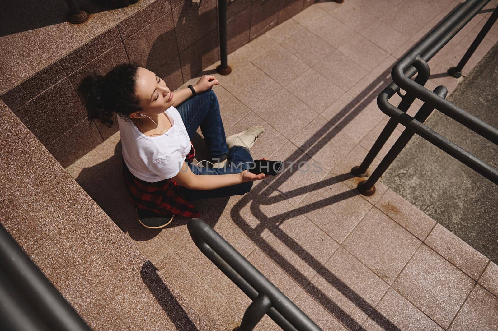 High angle view of a woman relaxing sitting on skateboard with headphones and smartphone in her hand and taking sun bathing outdoors