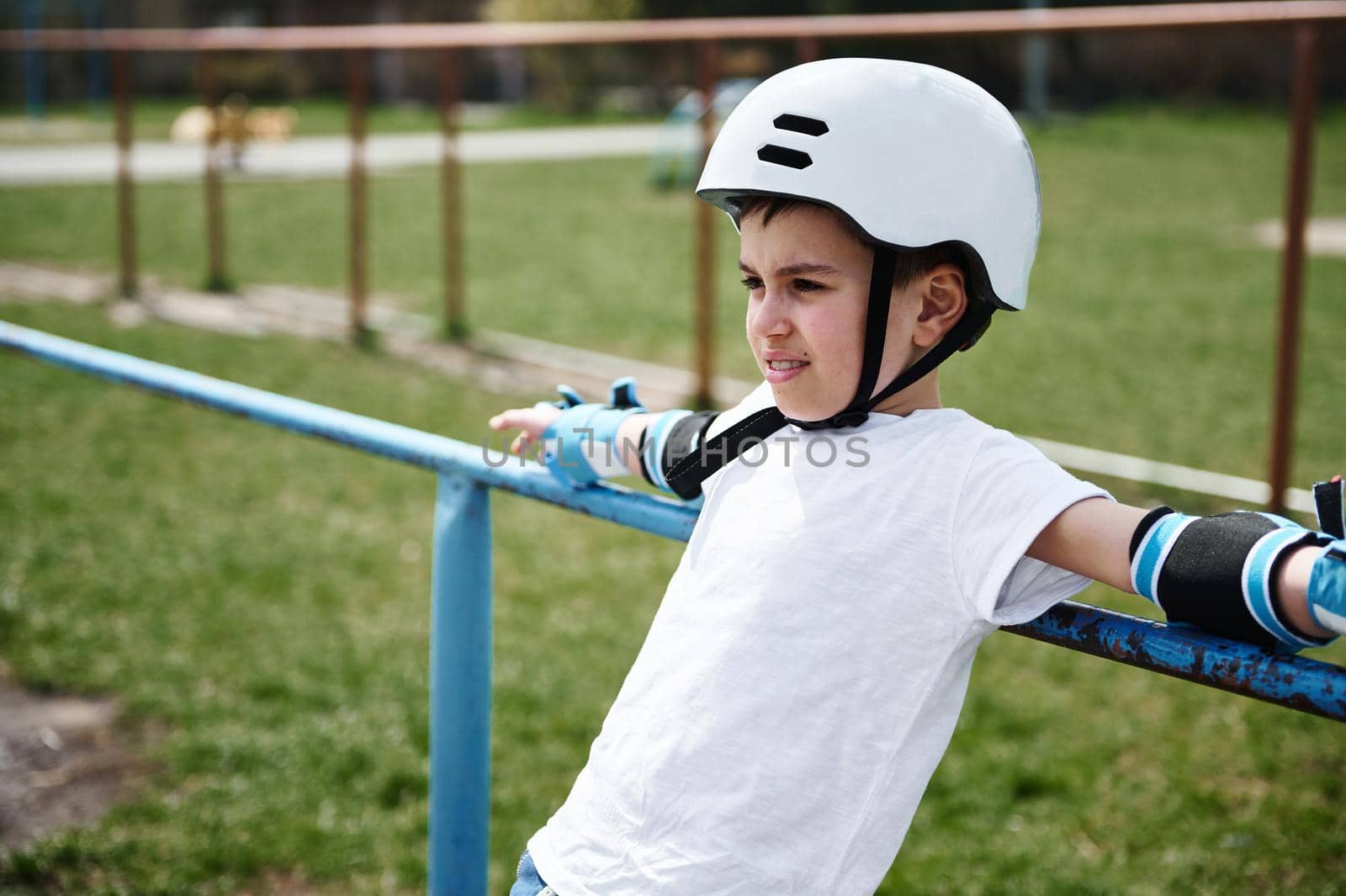 Handsome boy in safety helmet and protective gear leaning against horizontal bar on playground outdoors. Closeup