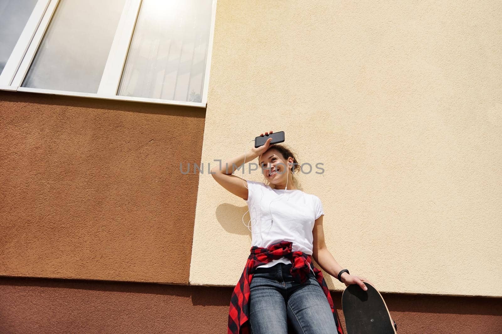 Beautiful hispanic woman in a white t-shirt and jeans posing with a smartphone and a skateboard in her hands against a colored wall on a beautiful sunny day